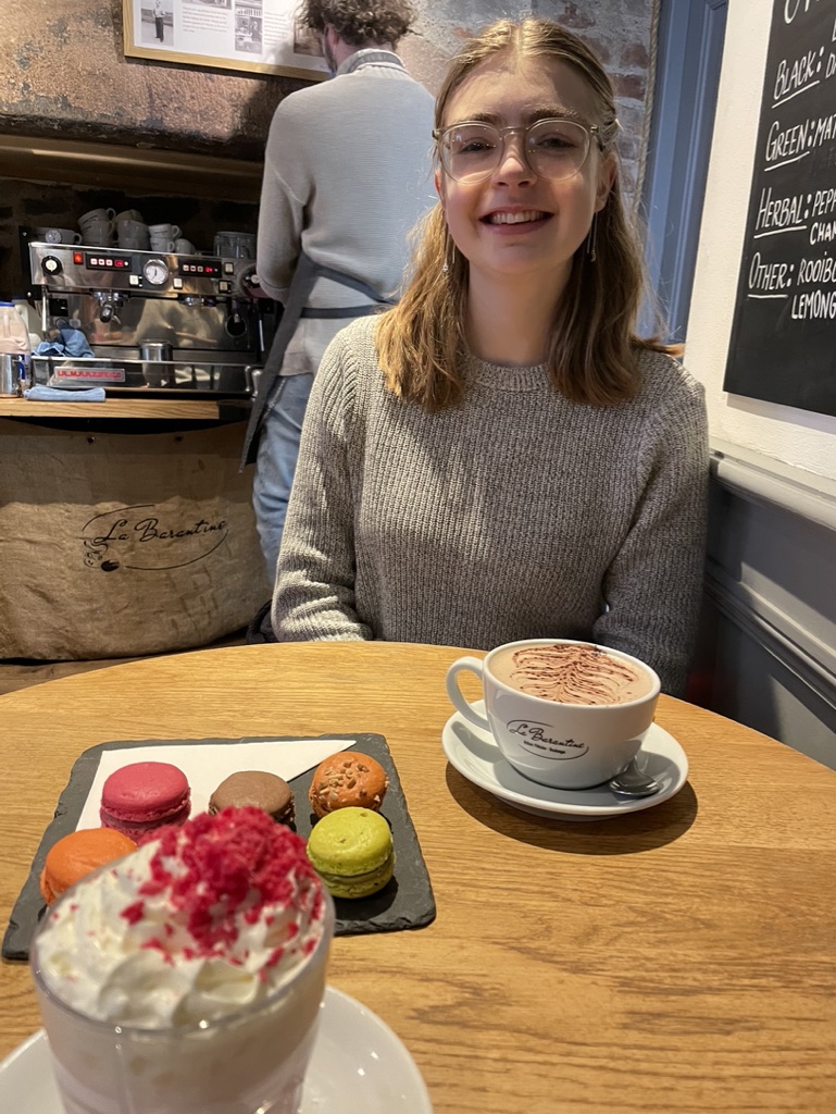 An image of Amelia, Amelia is sat in a  cafe wearing a grey jumper