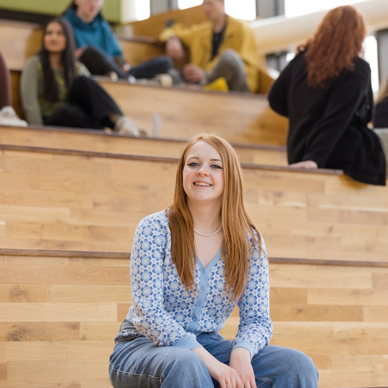 Biology student Briony in a pale blue top and blue jeans, sat on wooden steps