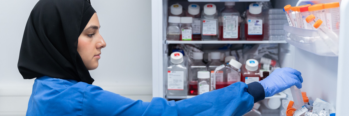 Student in a blue lab coat and a black head scarf placing samples into a fridge