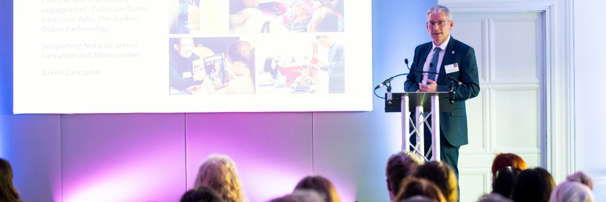 Professor Andy Schofield stands on a stand in front of a screen and the audience