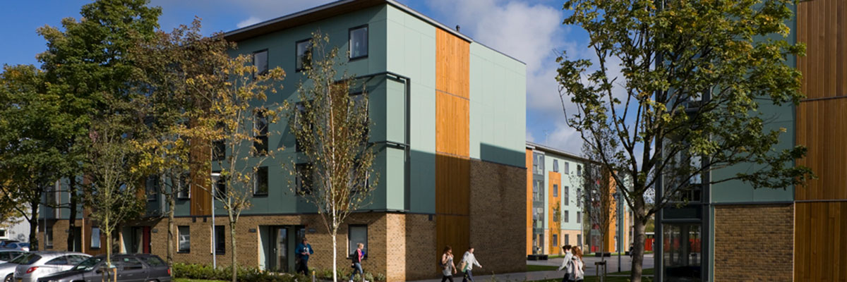 Green with wooden panelling accommodation building with parked cars and trees in front.