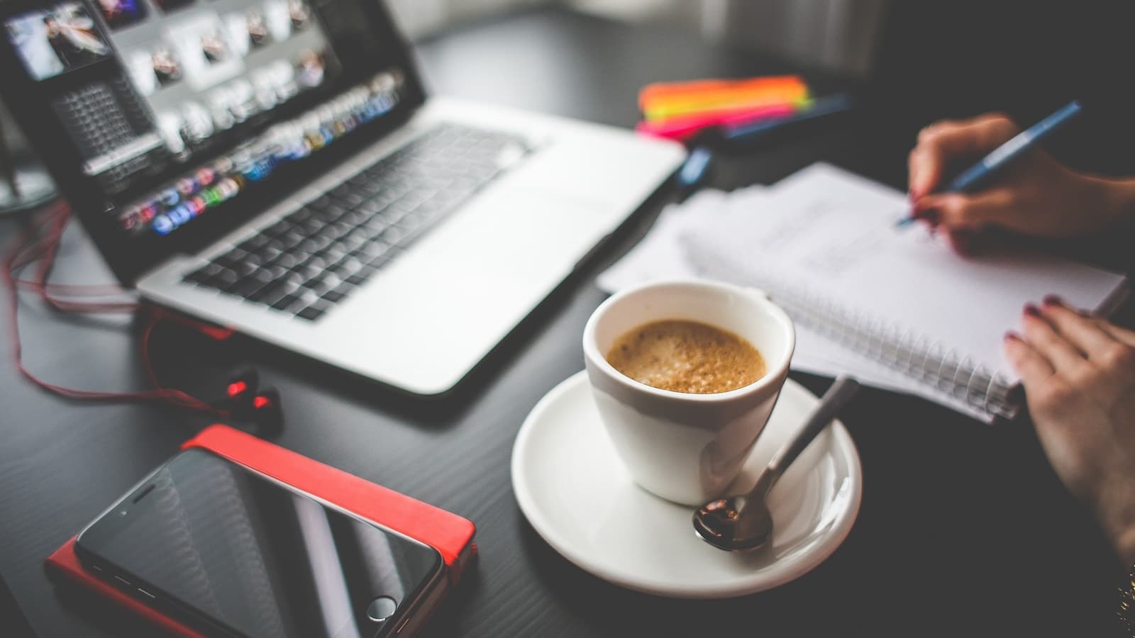 Workspace with cup of coffee and mobile phone in foreground, and the hands of a person writing on a notepad and a laptop in the background.