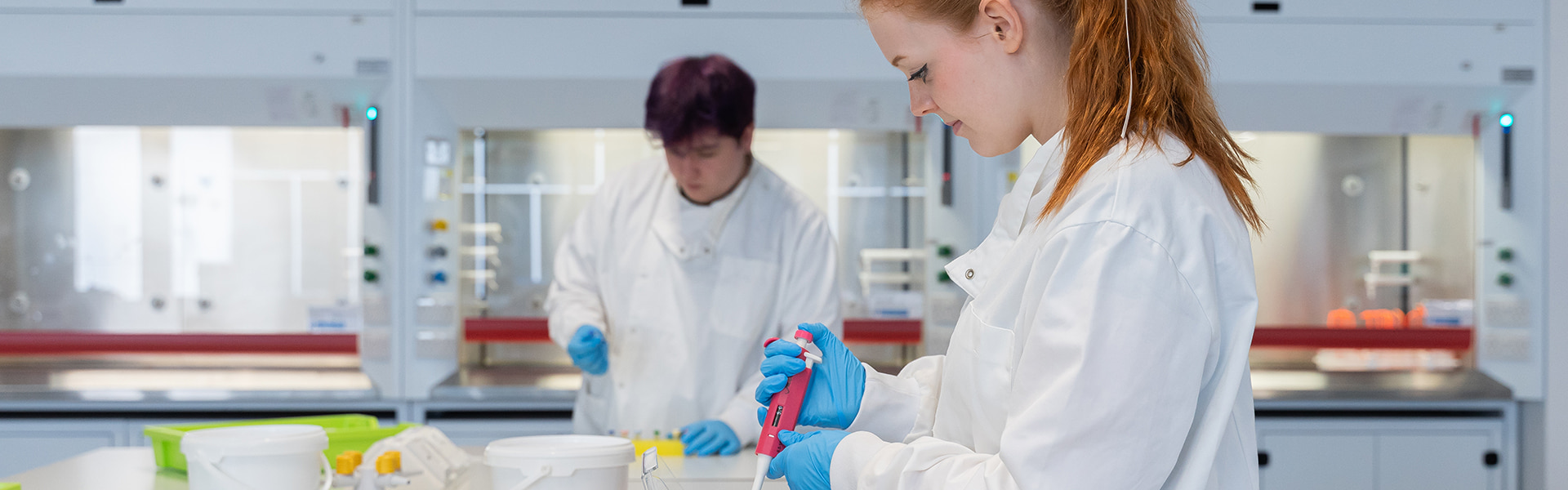 Two students in white coats and blue gloves working in a clinical laboratory