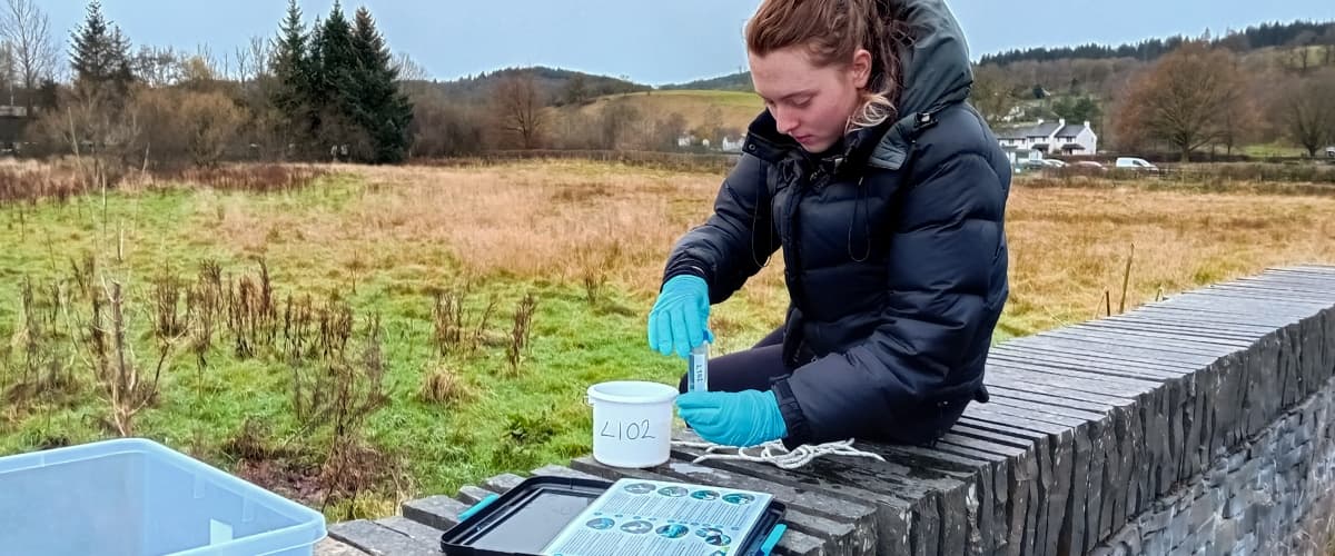 A person in an anorak takes a water sample in Hawkshead