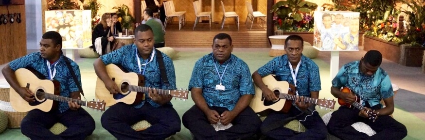 5 musicians of Fijian heritage, wearing patterned blue shirts and black trousers sitting cross legged.  3 have guitars, 1 has a ukelele