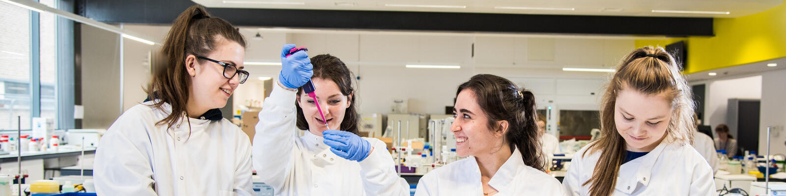 Three students in a lab wearing white lab coats. Student in middle is holding a pink pipette. 