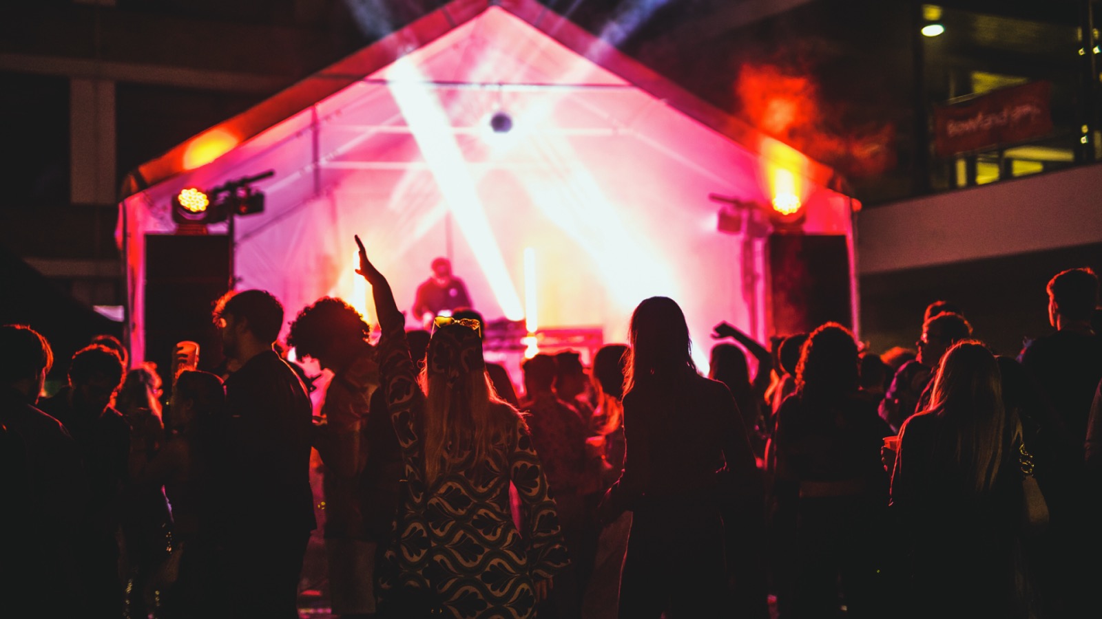 Students are silhouetted as they dance in front of a large tent housing a DJ and bright pink and red flashing lights.