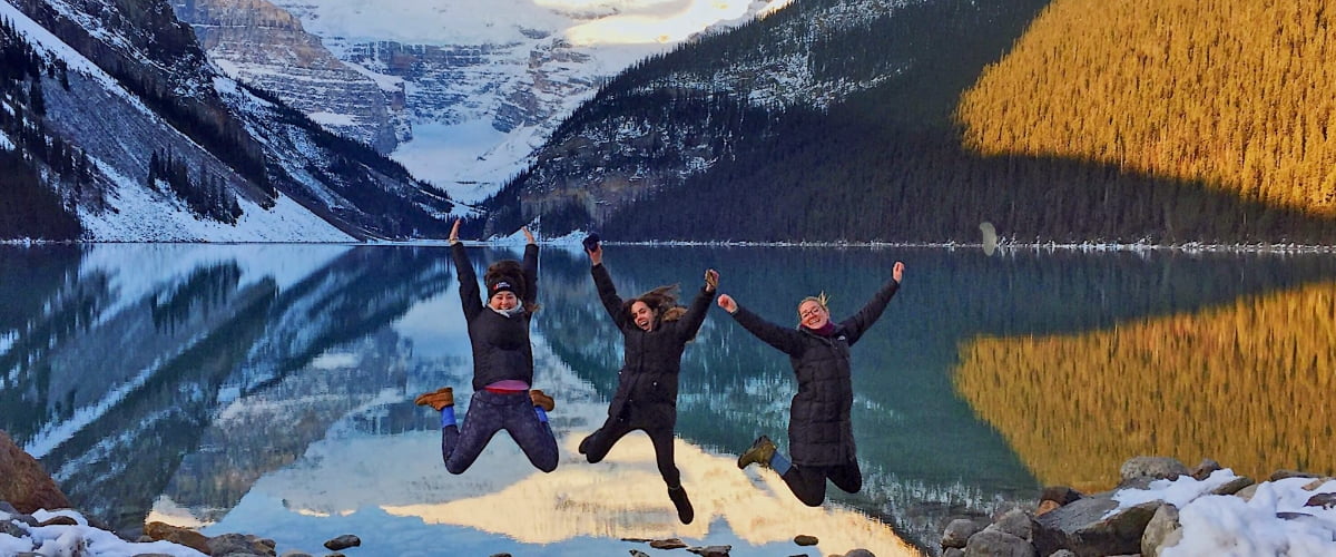 Three students leap into the air in front of a smooth lake, whose surface reflects the snow-covered mountains in the distance.