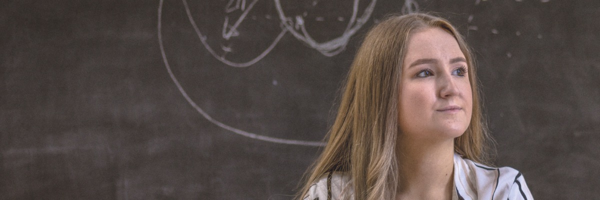 A student sits in front of a blackboard and stares off-camera right into the middle distance.